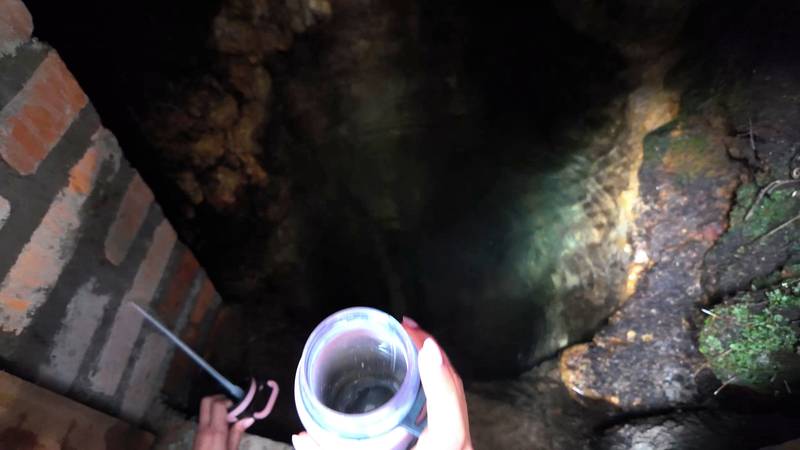 A hand lifting a glass jar of clear water out of a spring inside a brick-lined hatch