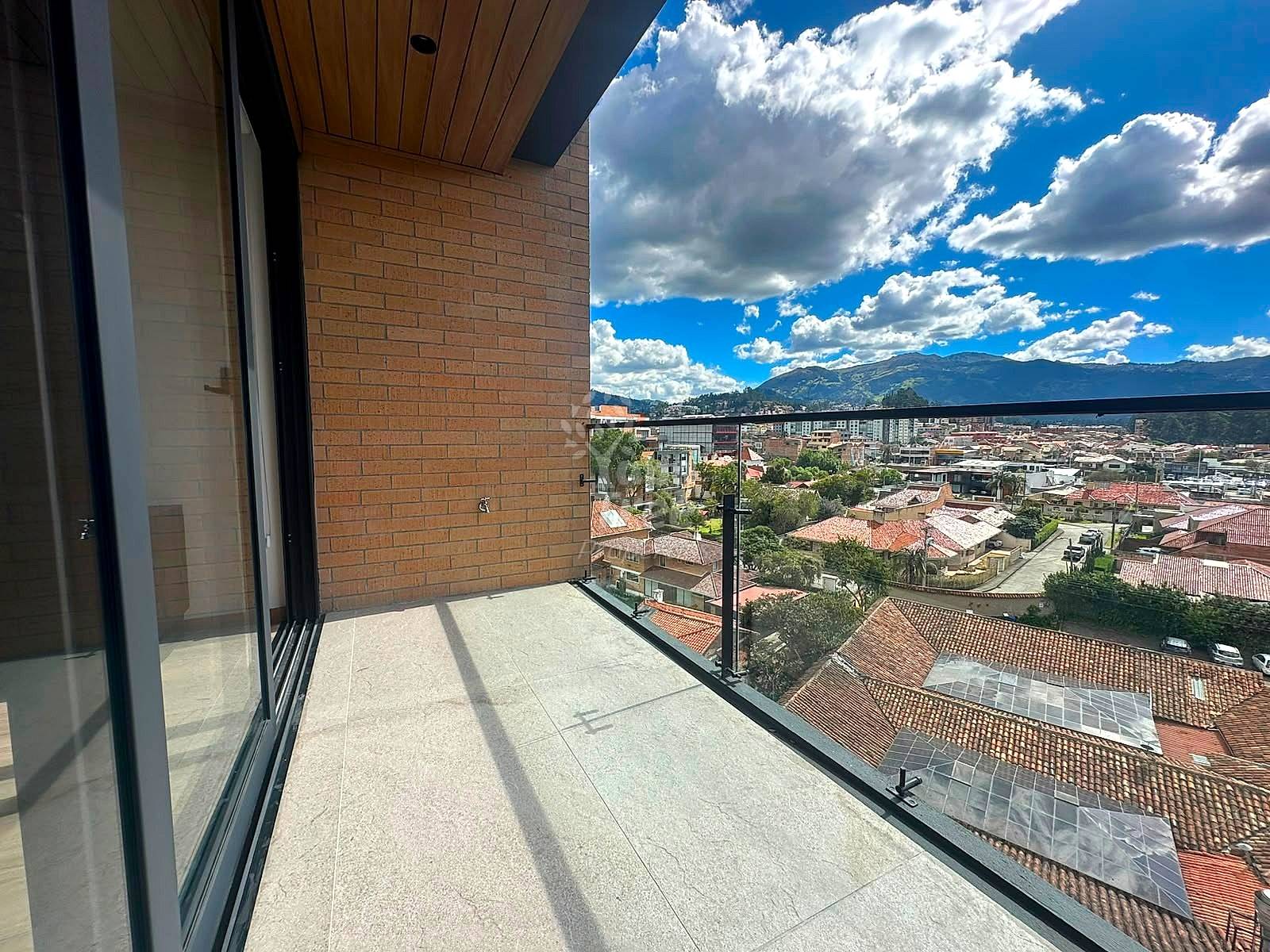 Balcony with gray tile flooring, brick wall, black glass railing, overlooking Cuenca cityscape with mountains and blue sky.