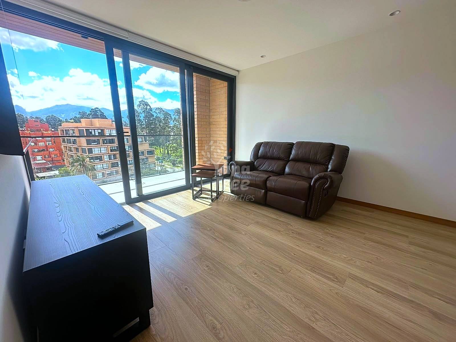 Living room with brown leather sofa, wooden flooring, flat-screen TV, and large glass doors opening to balcony with mountain views.