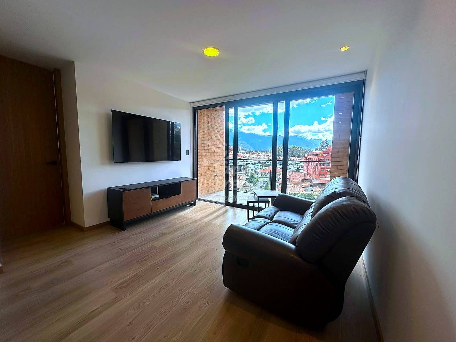 Living room with black leather sofa, wall-mounted TV, wooden floor, and floor-to-ceiling windows overlooking Cuenca valley and mountains.