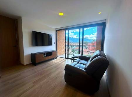 Living room with black leather sofa, wall-mounted TV, wooden floor, and floor-to-ceiling windows overlooking Cuenca valley and mountains.