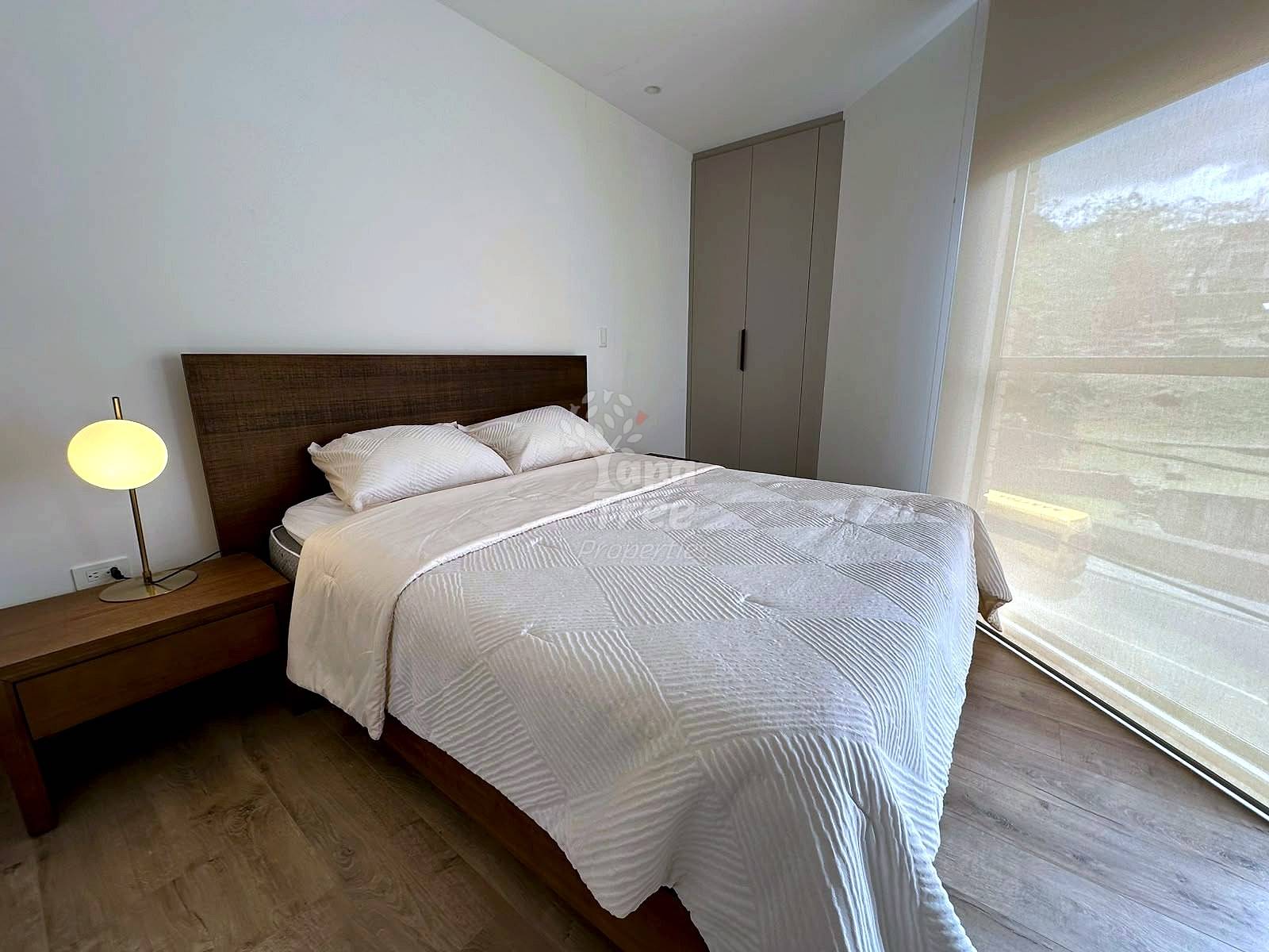 Bedroom with dark wood bed frame, white bedding, two pillows, wooden nightstand with yellow lamp, gray wardrobe, and window with natural light.