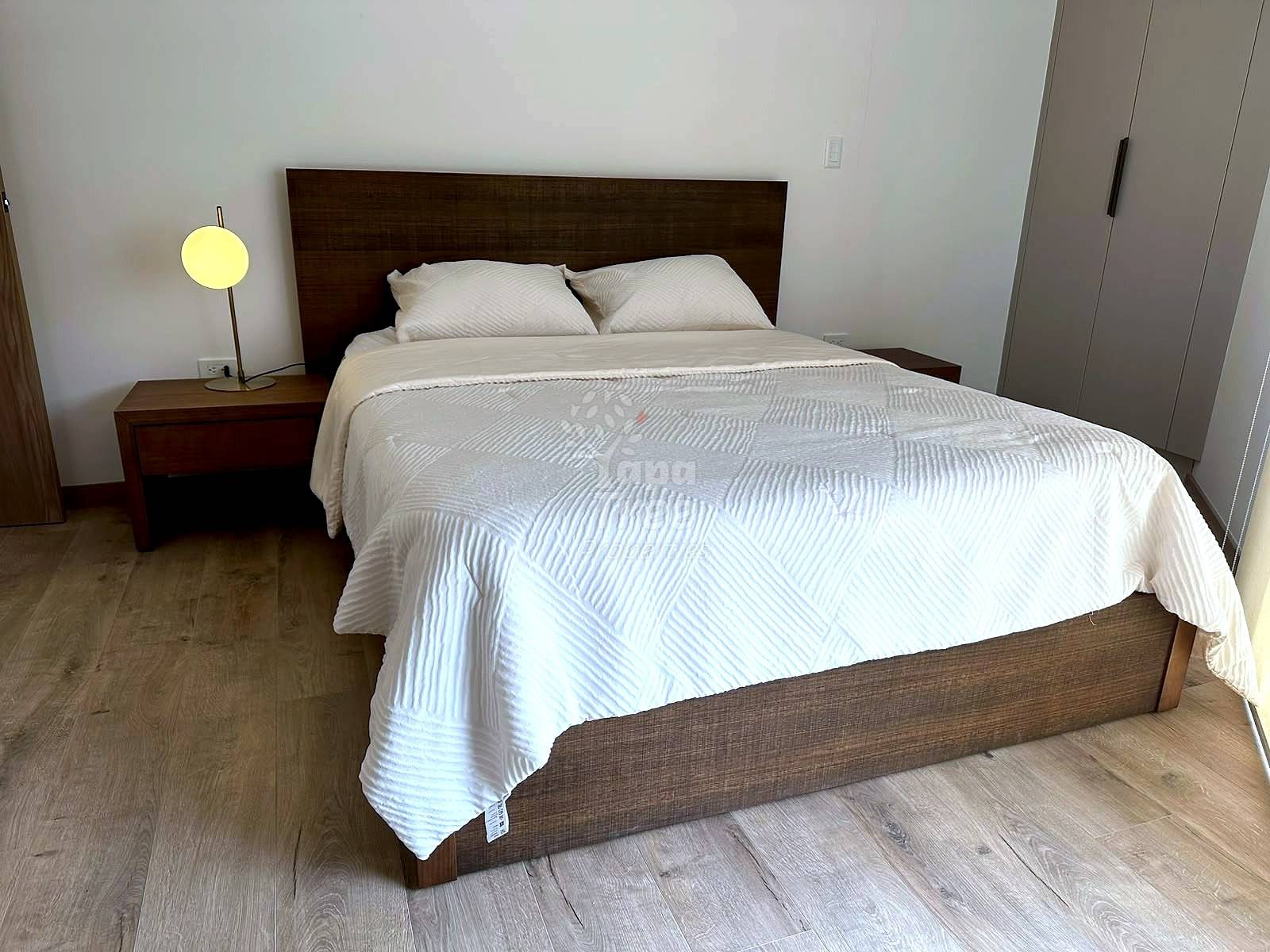 Bedroom with queen bed, dark wood headboard, cream bedding, wooden nightstand with yellow lamp, and light wood flooring.