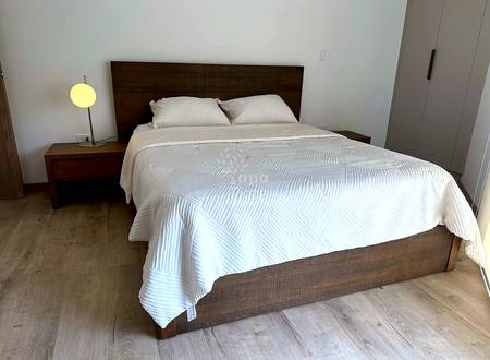 Bedroom with queen bed, dark wood headboard, cream bedding, wooden nightstand with yellow lamp, and light wood flooring.