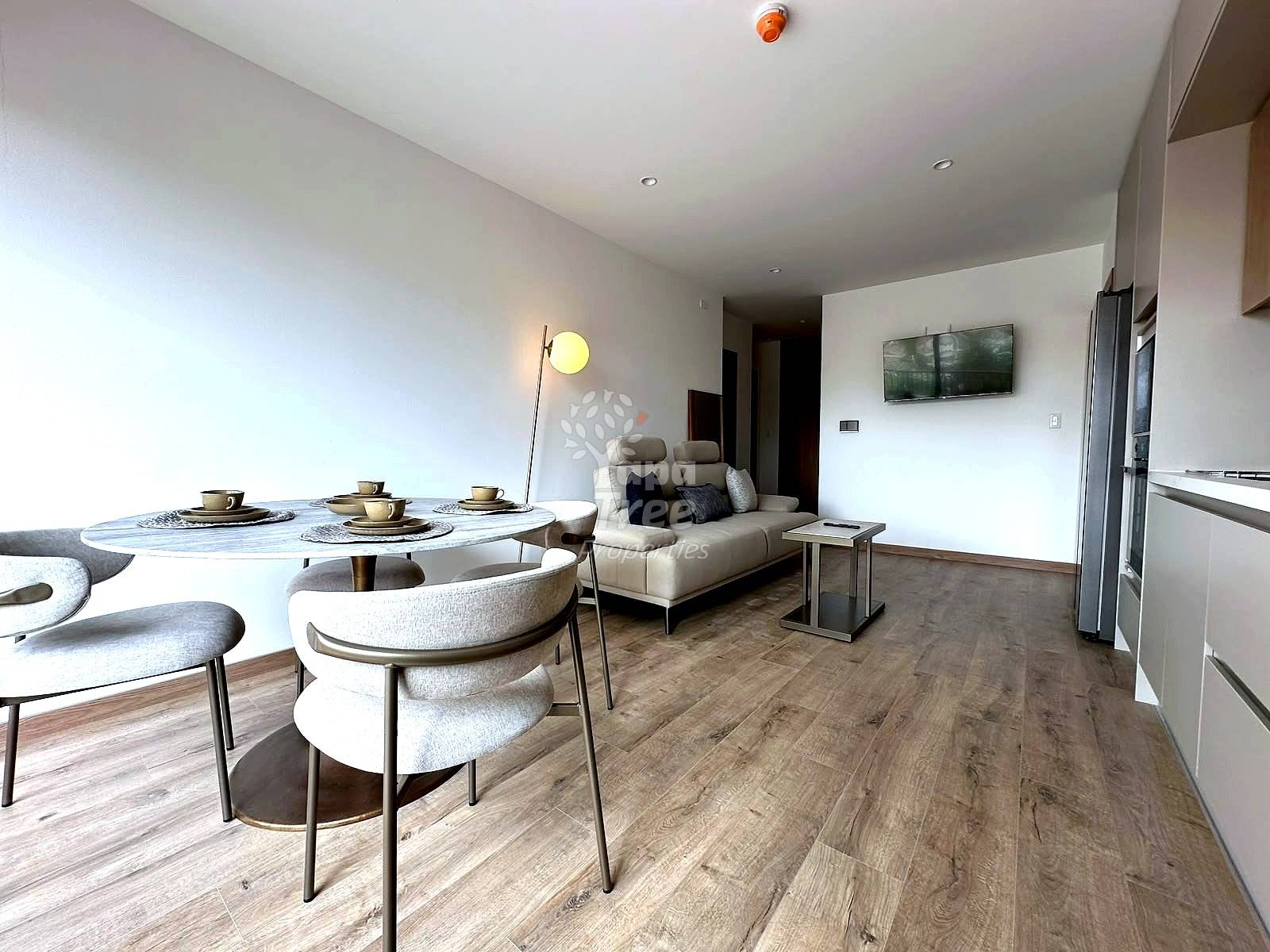 Open-plan living and dining area with wood-look flooring, round white table with cream chairs, beige sofa, floor lamp, and kitchen visible on right.
