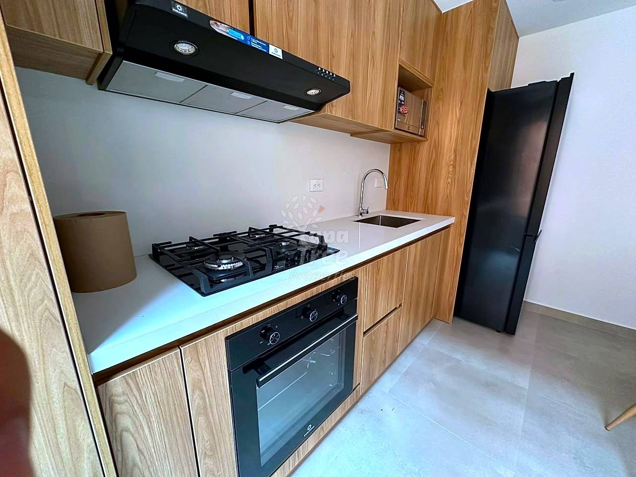 Modern cocina with wood cabinetry, white countertop, gas cooktop, built-in oven, black range hood, and stainless steel sink under bright lighting.