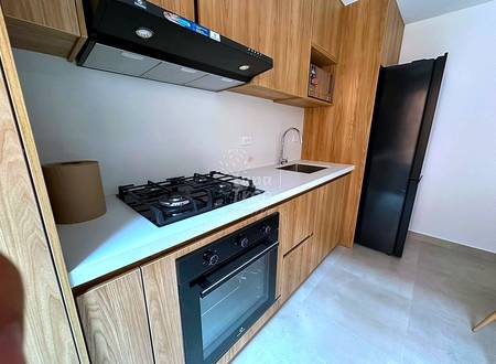 Modern cocina with wood cabinetry, white countertop, gas cooktop, built-in oven, black range hood, and stainless steel sink under bright lighting.