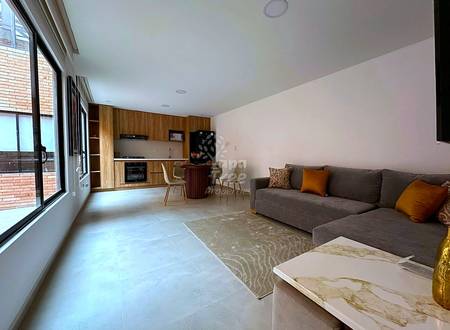 Open-plan living room with gray sectional sofa, wooden kitchen cabinets, dining area, large windows, and polished concrete flooring.