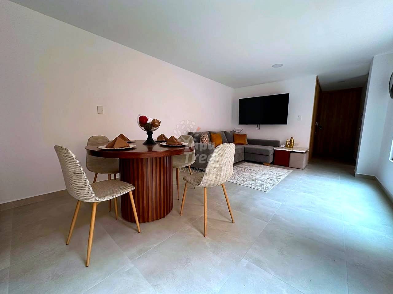 Open living and dining area with round wooden table, beige chairs, gray sectional sofa, wall-mounted TV, and light tile flooring.