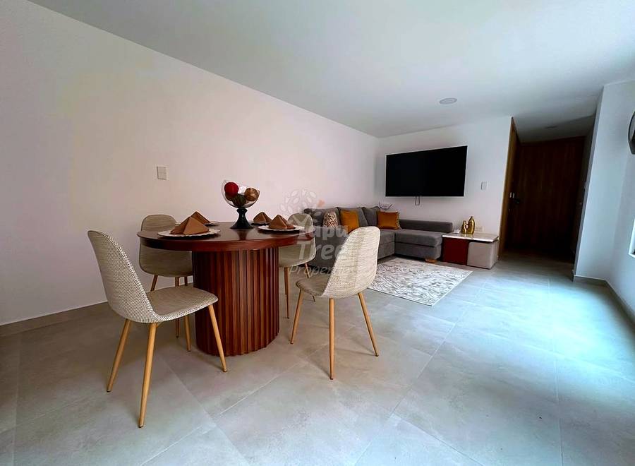Open living and dining area with round wooden table, beige chairs, gray sectional sofa, wall-mounted TV, and light tile flooring.