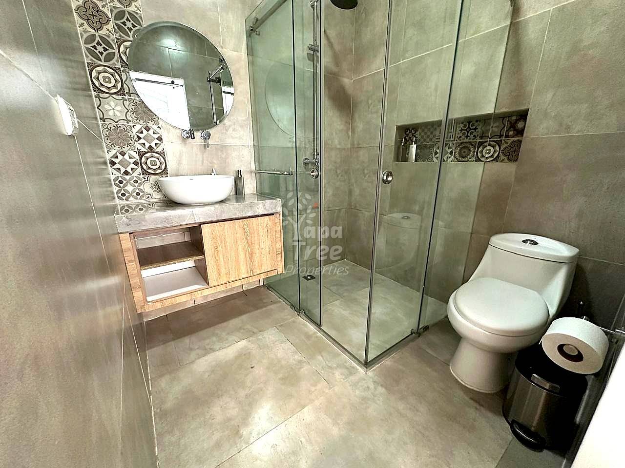Bathroom with gray tile walls, white round sink on wood vanity, round mirror, glass shower enclosure, and white toilet with trash bin.