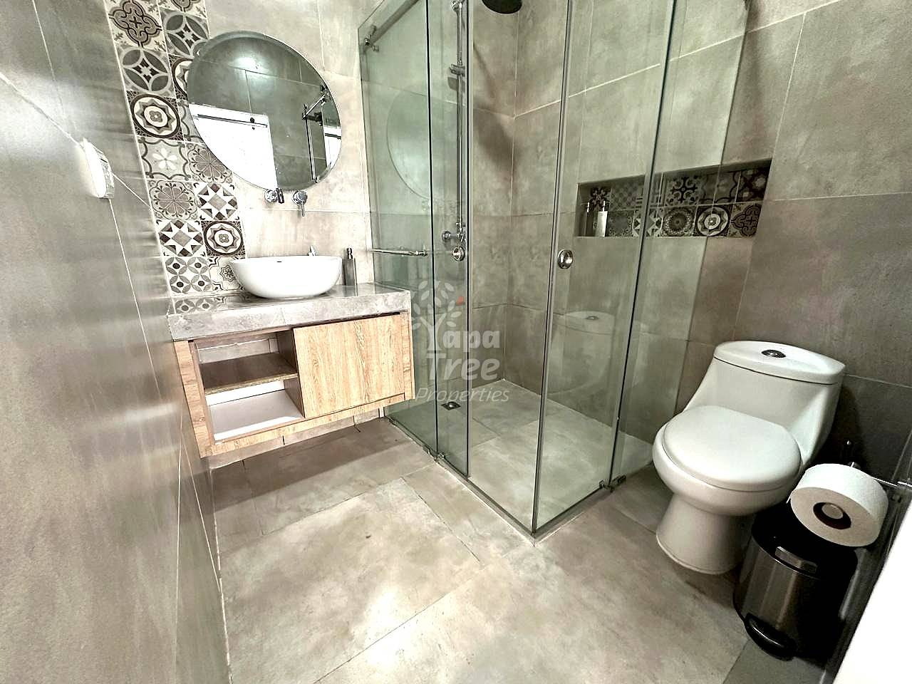 Bathroom with gray tile walls, white round sink on wood vanity, round mirror, glass shower enclosure, and white toilet with trash bin.