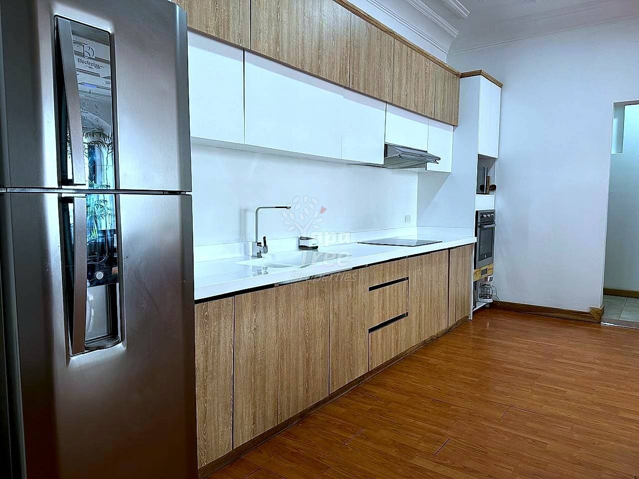 Modern cocina with wood cabinetry, white countertop, stainless steel refrigerator, gas cooktop, and wood plank flooring under bright overhead lighting.