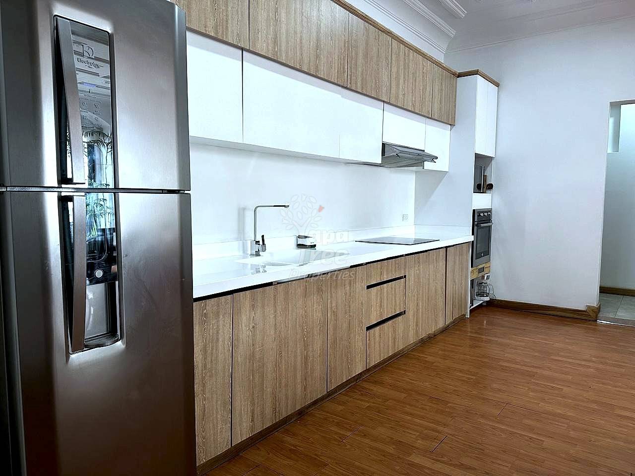 Modern cocina with wood cabinetry, white countertop, stainless steel refrigerator, gas cooktop, and wood plank flooring under bright overhead lighting.