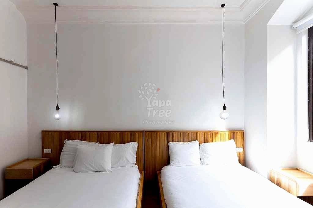 Bedroom with two twin beds, wooden headboards, white linens, pendant lights, and natural window light from the right.
