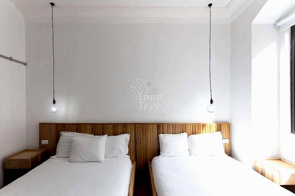 Bedroom with two twin beds, wooden headboards, white linens, pendant lights, and natural window light from the right.
