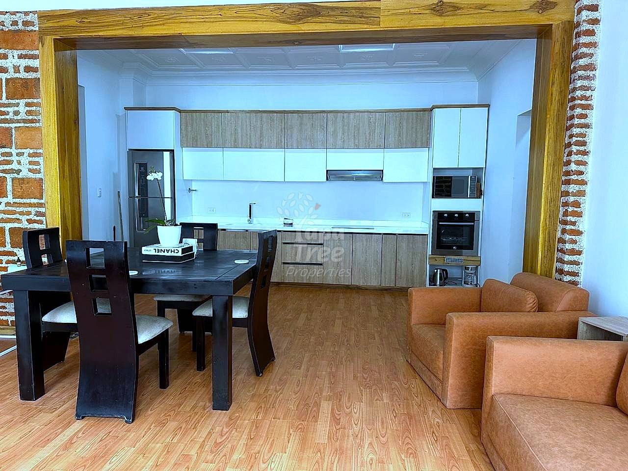 Open-concept departamento interior showing wood-grain kitchen cabinets, black dining table with chairs, and tan leather sofa on laminate flooring.