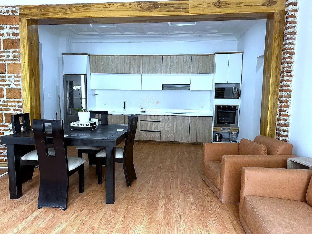 Open-concept departamento interior showing wood-grain kitchen cabinets, black dining table with chairs, and tan leather sofa on laminate flooring.