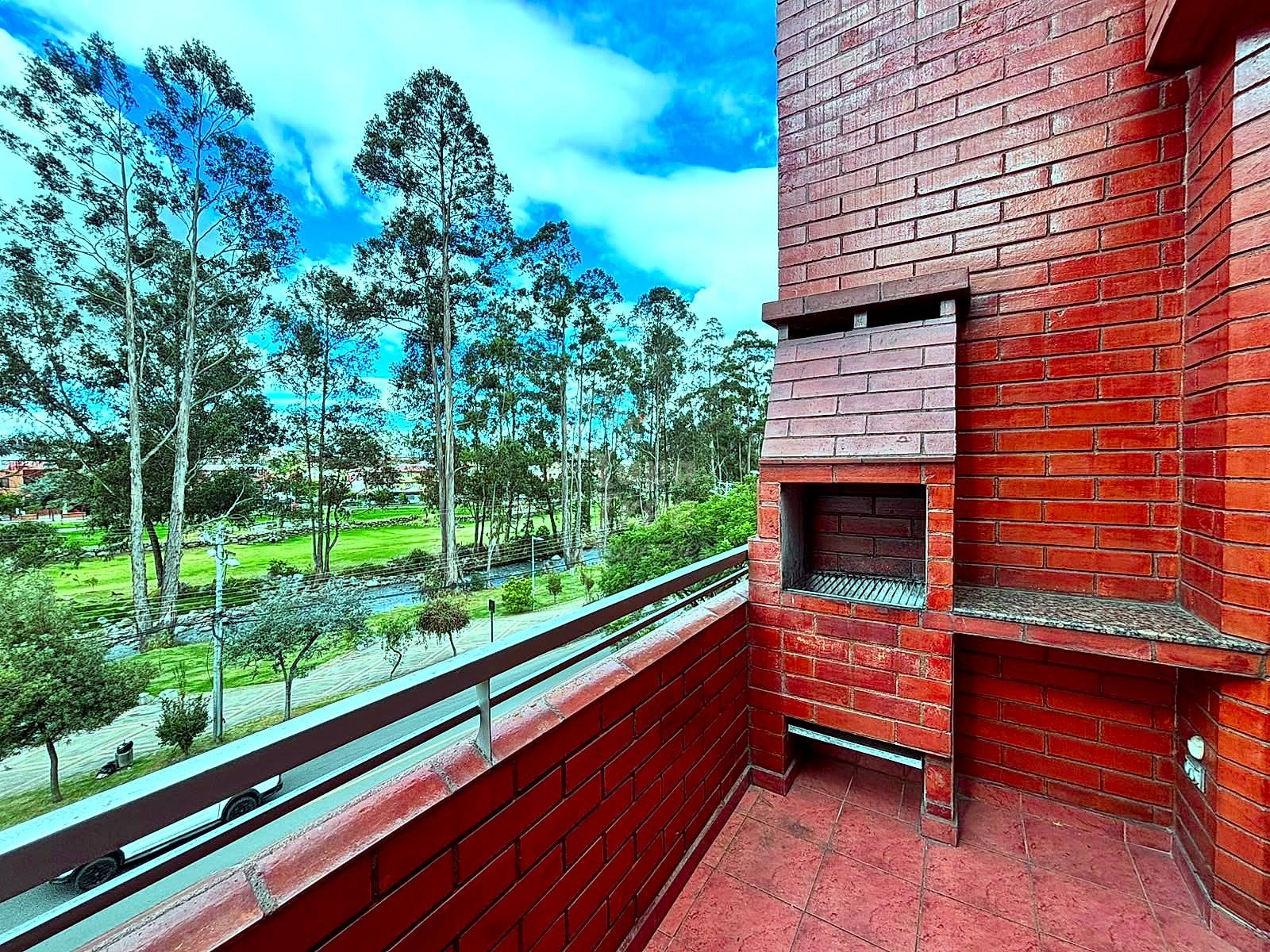 Red brick balcony with metal railing and built-in grill, overlooking park with tall trees and green lawn under blue sky.