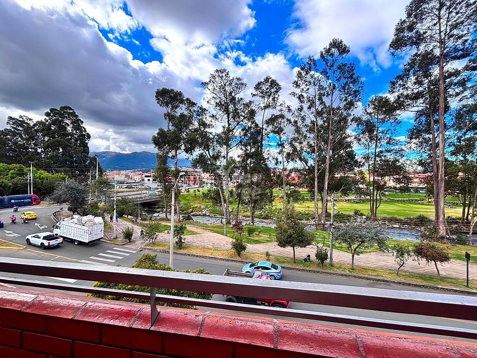 Elevated balcony view with metal railing overlooking park with tall eucalyptus trees, green lawn, paved pathways, streets, and distant Cuenca cityscape under cloudy sky.