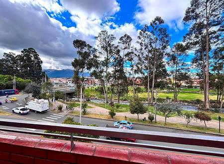 Elevated balcony view with metal railing overlooking park with tall eucalyptus trees, green lawn, paved pathways, streets, and distant Cuenca cityscape under cloudy sky.