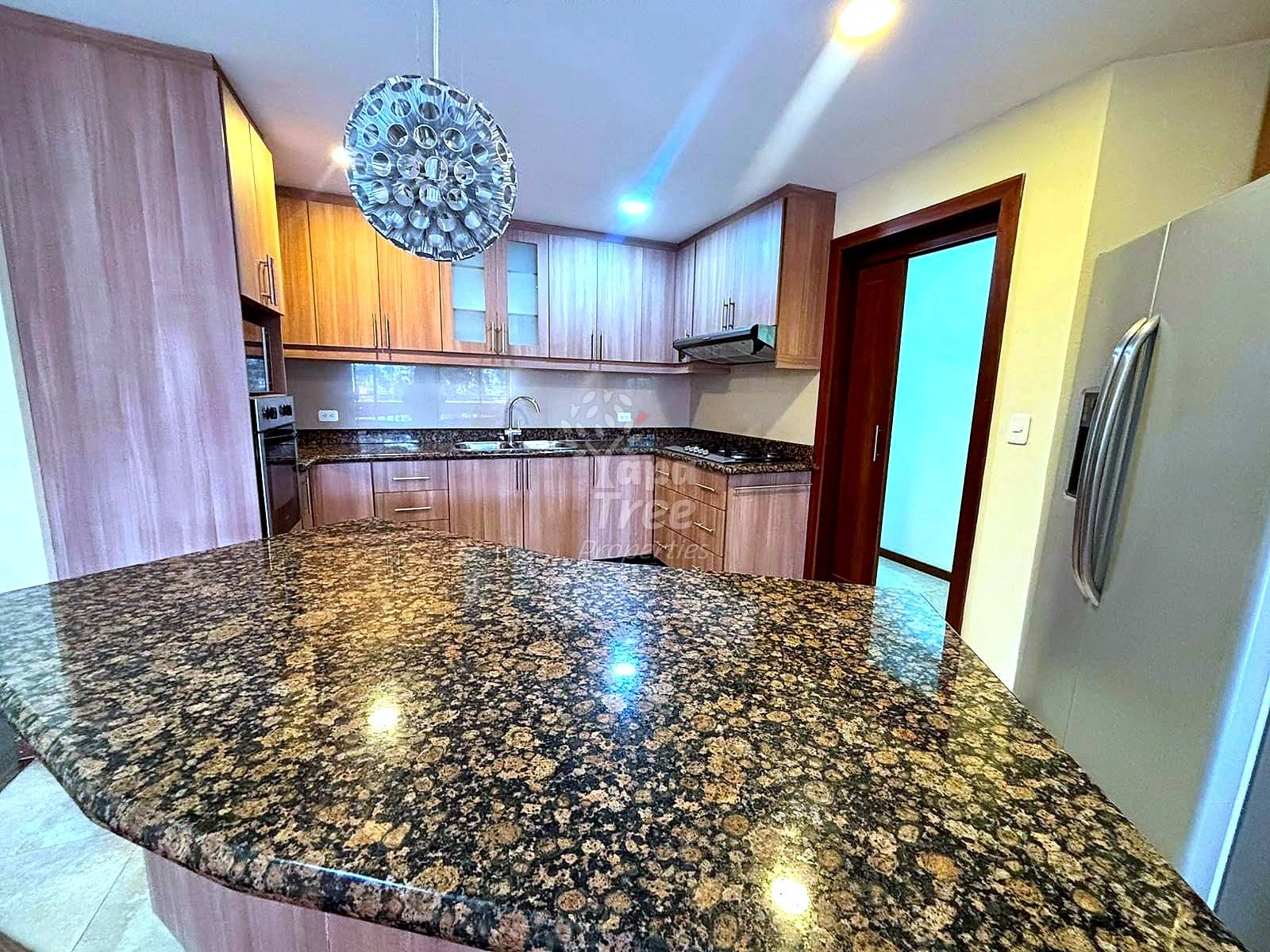 Modern cocina with wooden cabinetry, granite countertop island, stainless steel refrigerator, pendant lighting, and cream-colored walls.