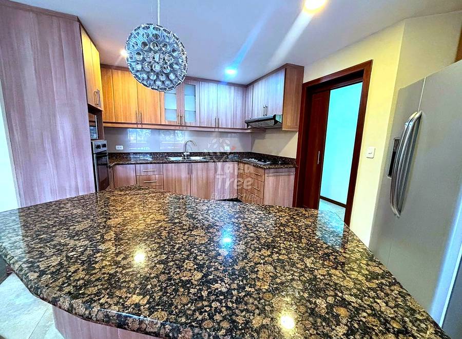 Modern cocina with wooden cabinetry, granite countertop island, stainless steel refrigerator, pendant lighting, and cream-colored walls.