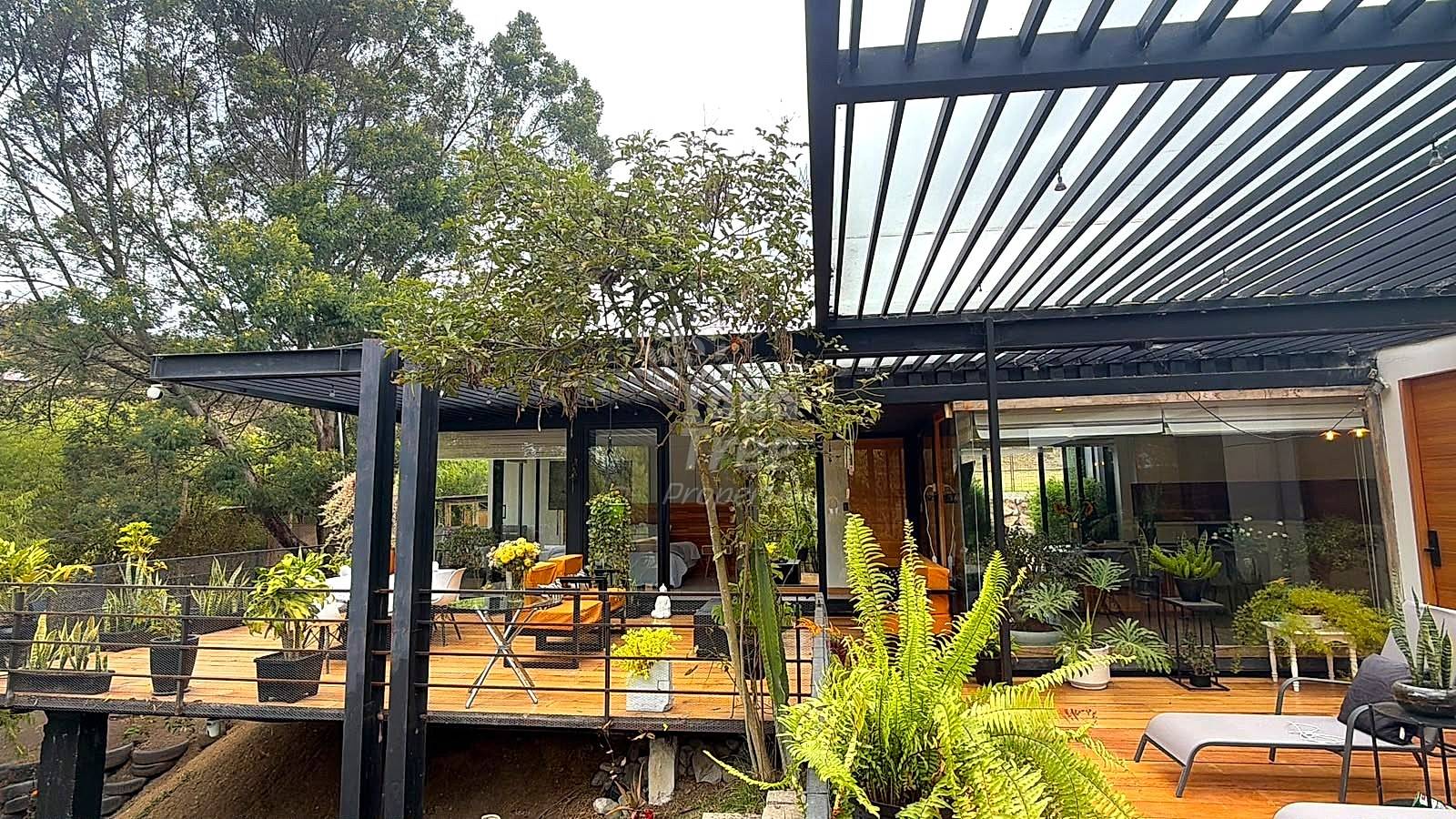 Modern deck with black metal pergola, wooden flooring, potted plants, and glass-walled interior space visible beyond.