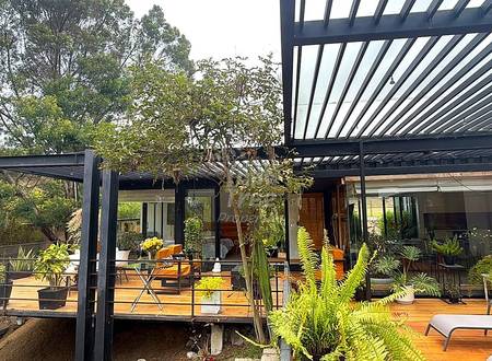 Modern deck with black metal pergola, wooden flooring, potted plants, and glass-walled interior space visible beyond.