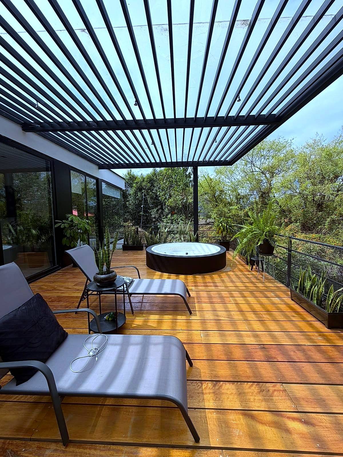 Covered terrace with wood decking, black pergola roof, lounge chairs, round hot tub, potted plants, and mountain views through railing.