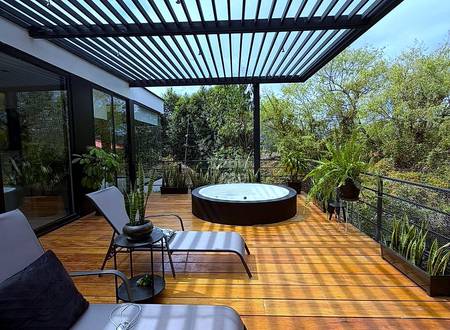 Covered terrace with wood decking, black pergola roof, lounge chairs, round hot tub, potted plants, and mountain views through railing.