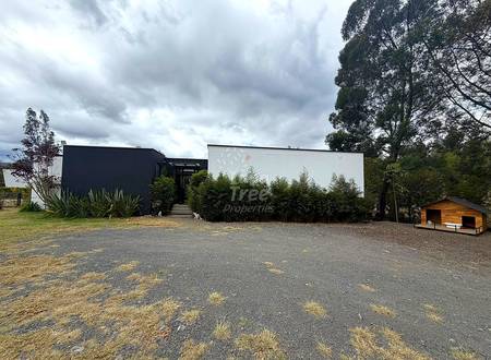 Modern residential property with black and white metal structures, gravel driveway, surrounding green vegetation, and wooden outbuilding under cloudy sky.