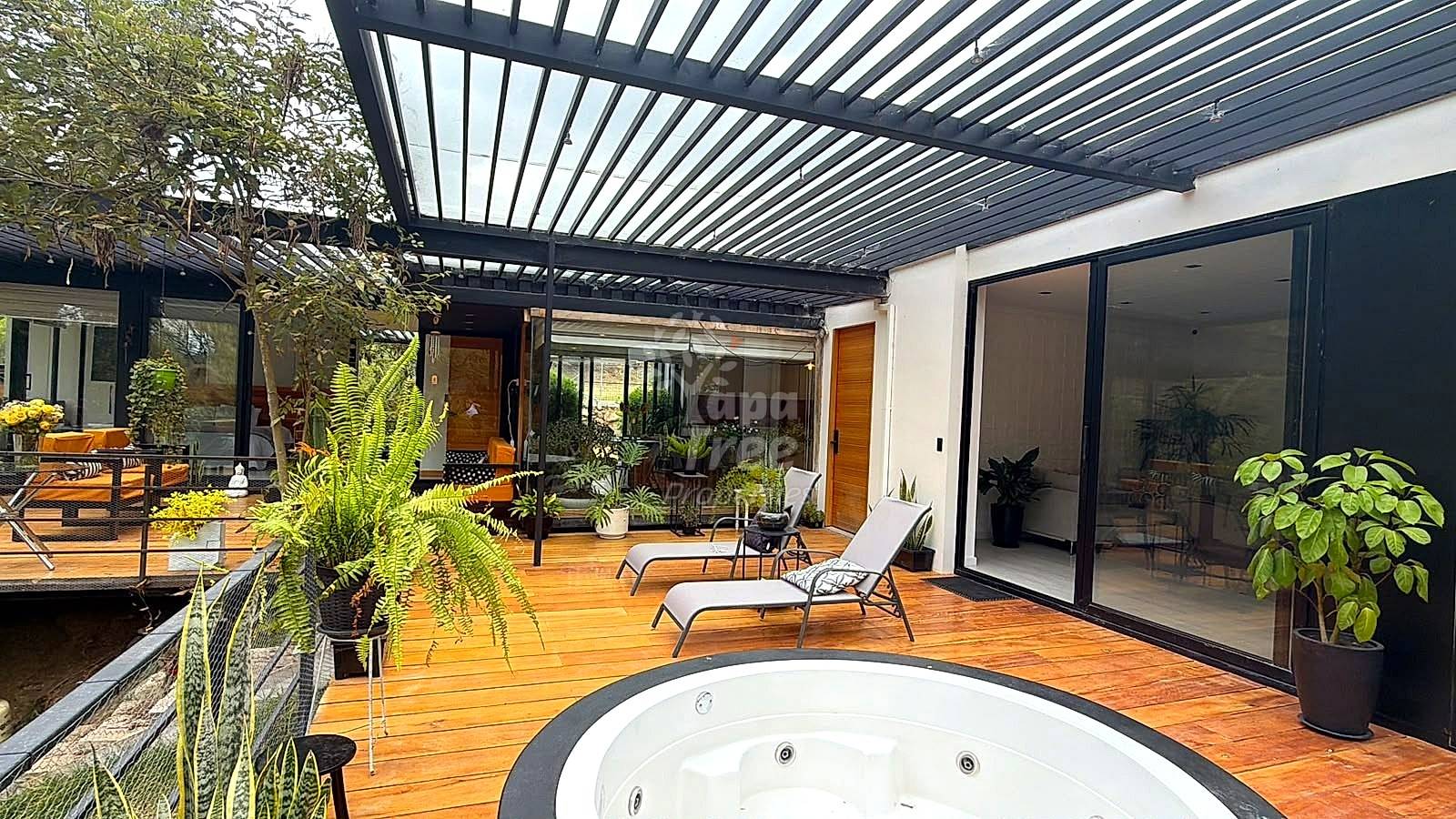 Outdoor patio with wooden deck, built-in hot tub, lounge chairs, potted plants, and slatted metal pergola overhead.
