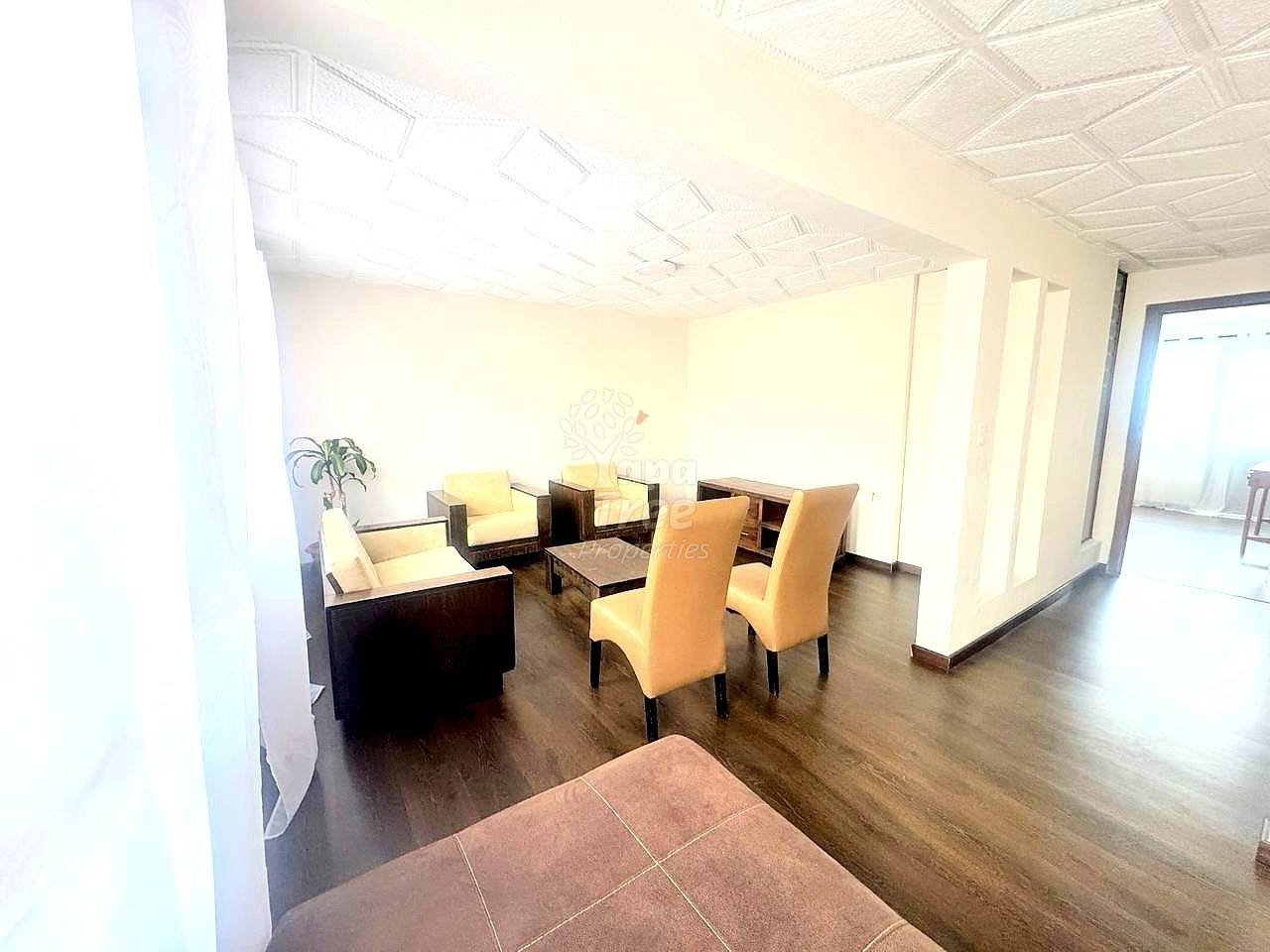 Open living area with wooden flooring, beige upholstered chairs, dark wood table, potted plant, and white drop ceiling tiles with recessed lighting.