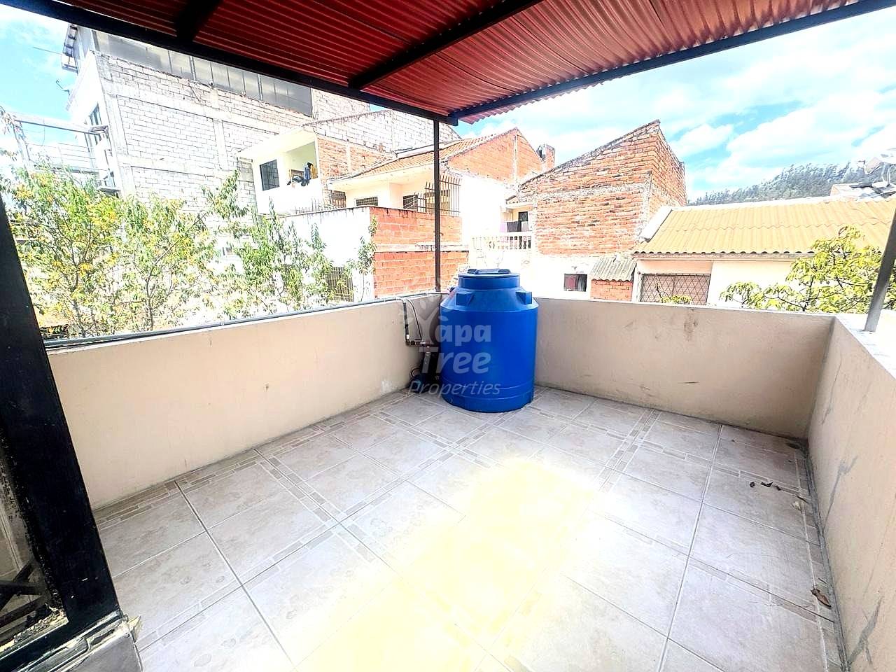Rooftop terrace with gray tile flooring, blue water storage tank, corrugated metal roof, and surrounding brick buildings in daylight.