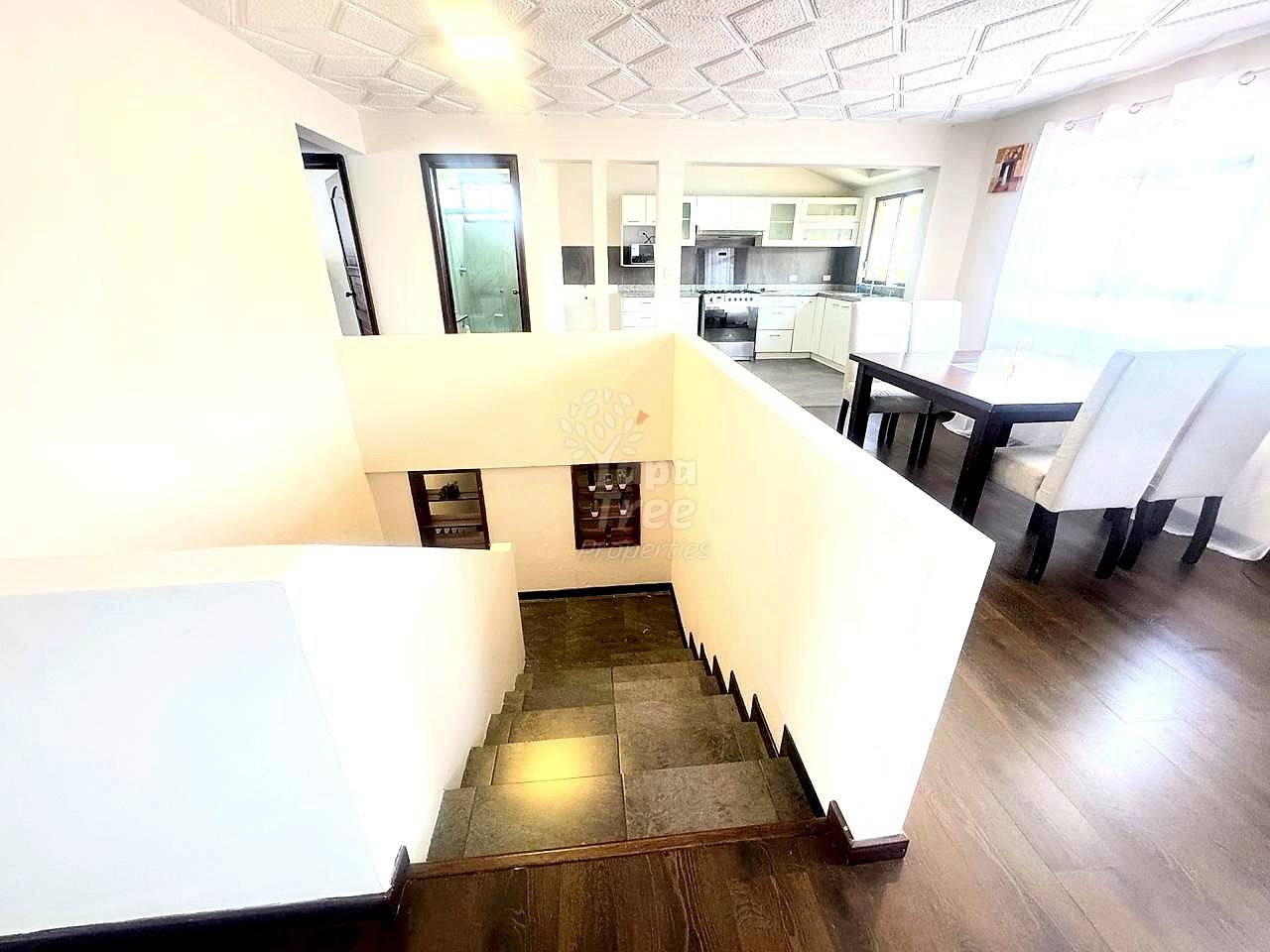 Staircase with tile steps descending to lower level, white walls, wooden handrail, open-plan departamento with white cocina and dining area visible above.