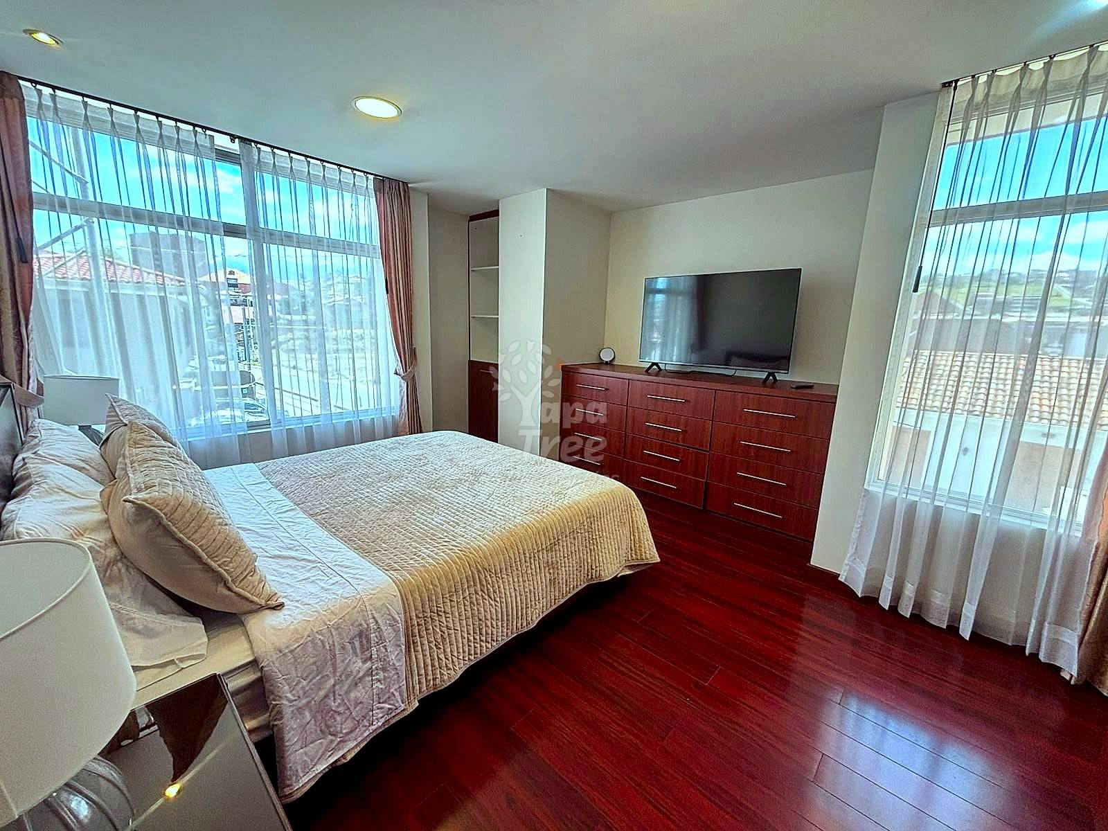 Bedroom with queen bed, dark wood dresser with flat-screen TV, dark wood flooring, and large windows with sheer curtains overlooking city views.