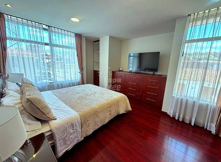 Bedroom with queen bed, dark wood dresser with flat-screen TV, dark wood flooring, and large windows with sheer curtains overlooking city views.