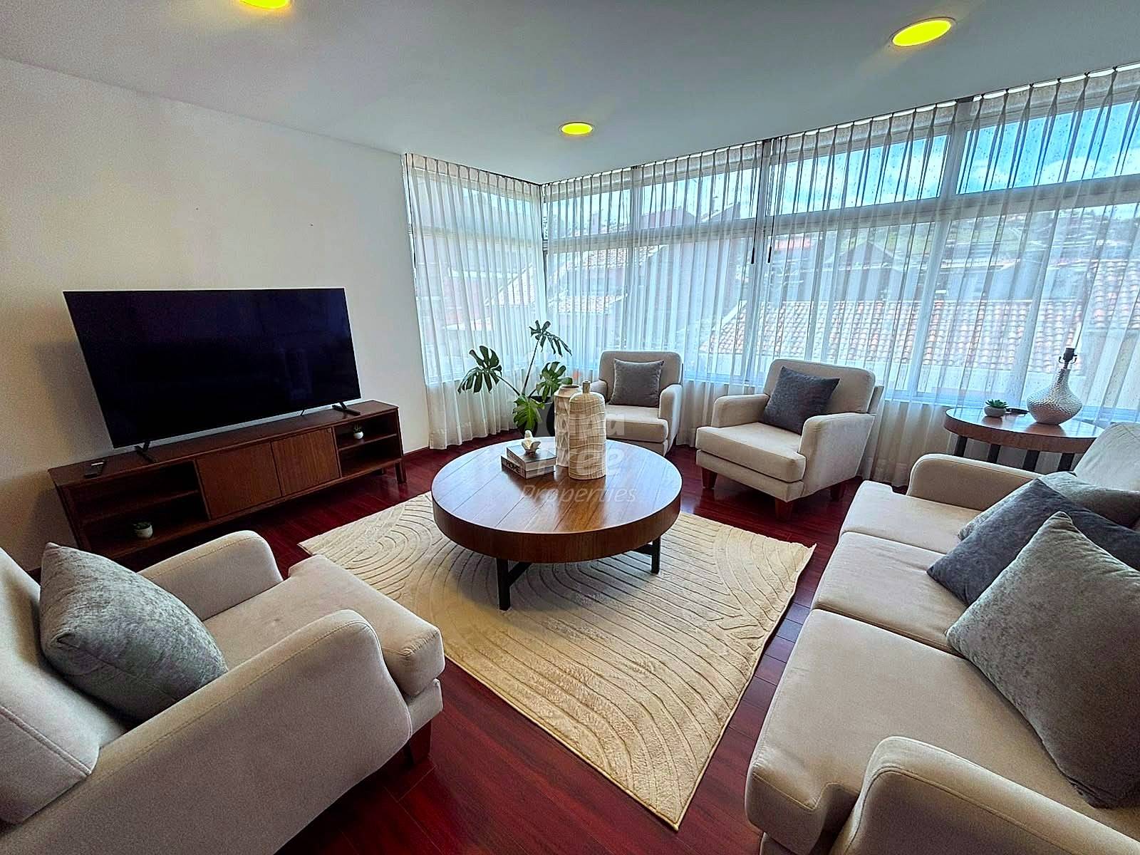 Living room with multiple beige upholstered chairs, round wood coffee table, TV on wood stand, large windows with vertical blinds, recessed ceiling lights, burgundy and neutral rug.