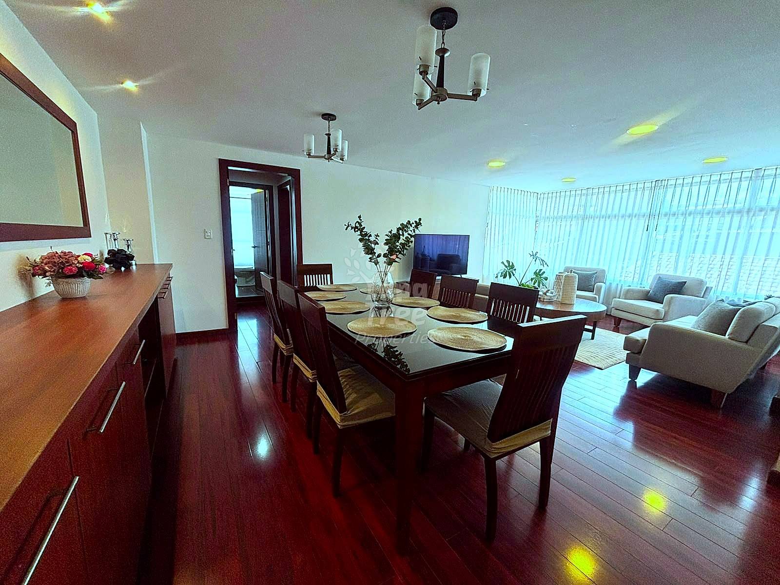 Open-concept dining and living room with dark wood dining table, six chairs, dark wood cabinetry, hardwood floors, recessed ceiling lights, and seating area with armchairs visible in background.