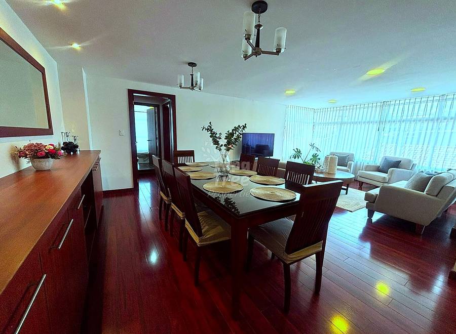Open-concept dining and living room with dark wood dining table, six chairs, dark wood cabinetry, hardwood floors, recessed ceiling lights, and seating area with armchairs visible in background.