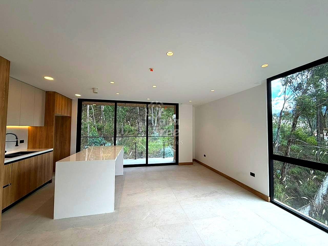 Open-plan living and kitchen area with white tile flooring, wood cabinetry, white island counter, recessed ceiling lights, and large black-framed windows overlooking green forest.