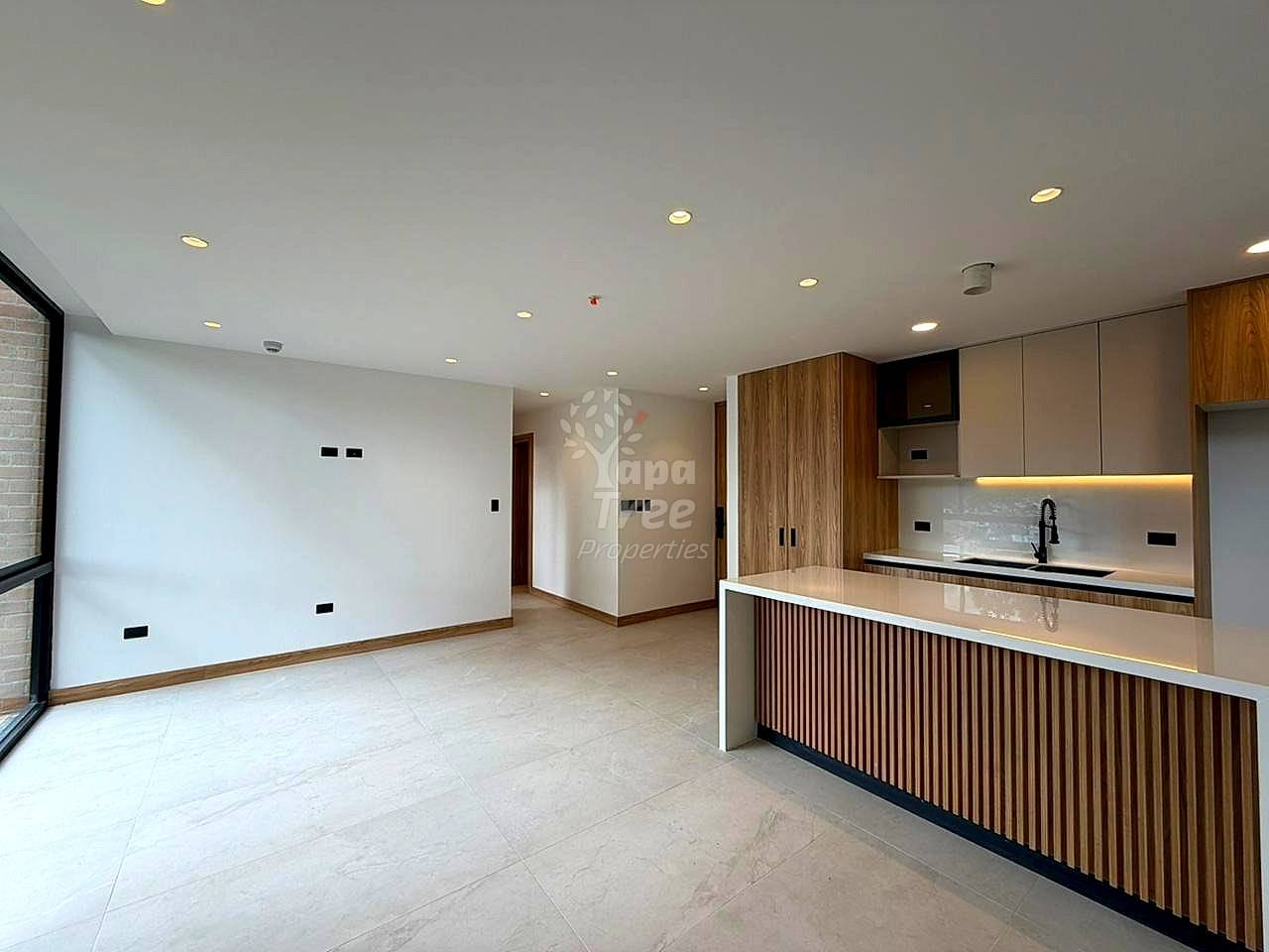 Modern open-concept departamento with white walls, recessed ceiling lights, wood-grain kitchen cabinetry, white countertop, and light tile flooring throughout.