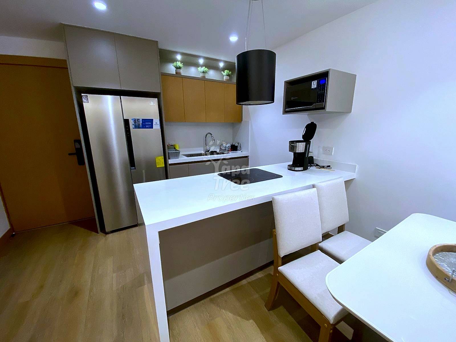 Modern kitchen with white island counter, stainless steel refrigerator, wood cabinetry, microwave, pendant light, and wooden dining table with cream chairs.