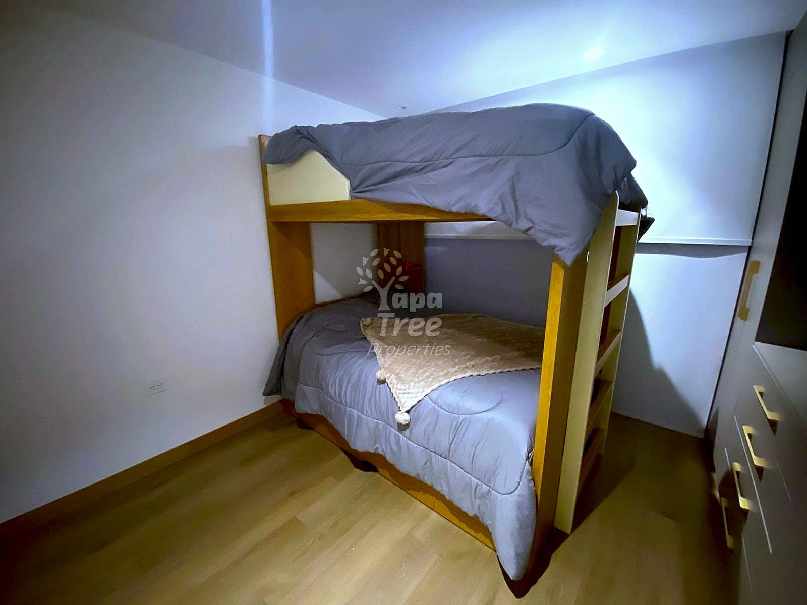 Small bedroom with wooden bunk bed frame, gray mattresses, and beige blankets under recessed ceiling lights.