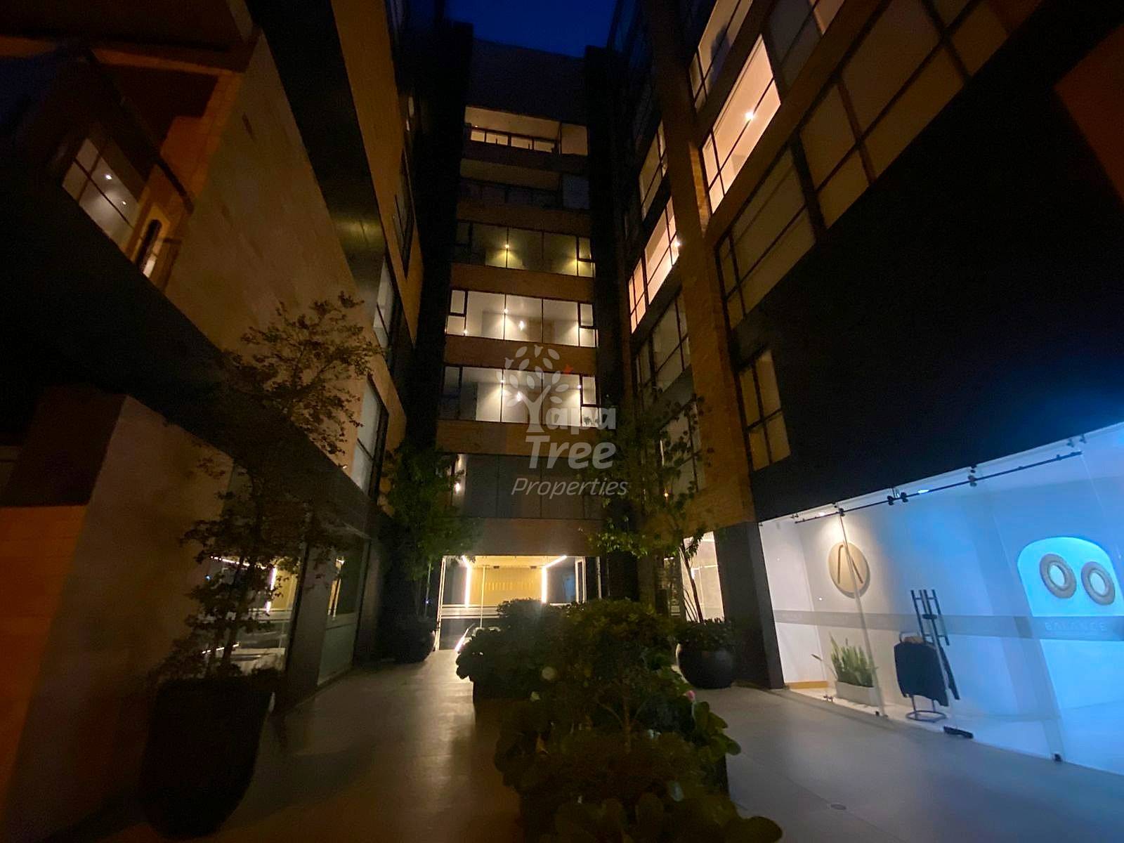 Night view of modern multi-story building entrance with lit windows, potted plants, and paved courtyard between surrounding structures.