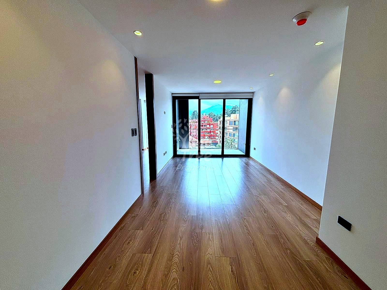 Empty room with wooden flooring, white walls, recessed ceiling lights, and glass sliding doors overlooking Cuenca cityscape with red brick buildings.