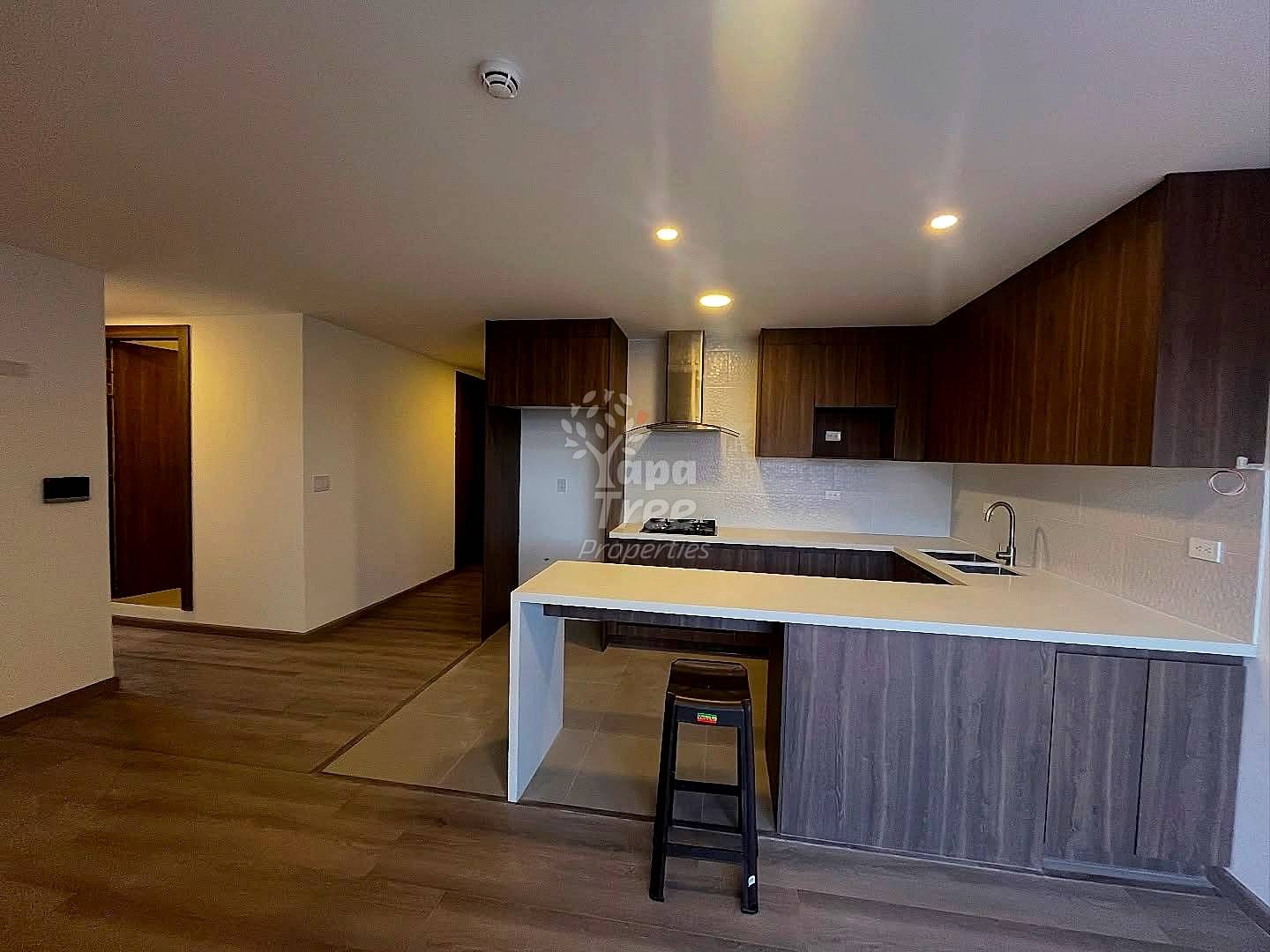 Cocina de concepto abierto con gabinetes de madera oscura, backsplash de azulejos blancos, estufa acero inoxidable, isla con mesón blanco, y luces embutidas en el techo.
