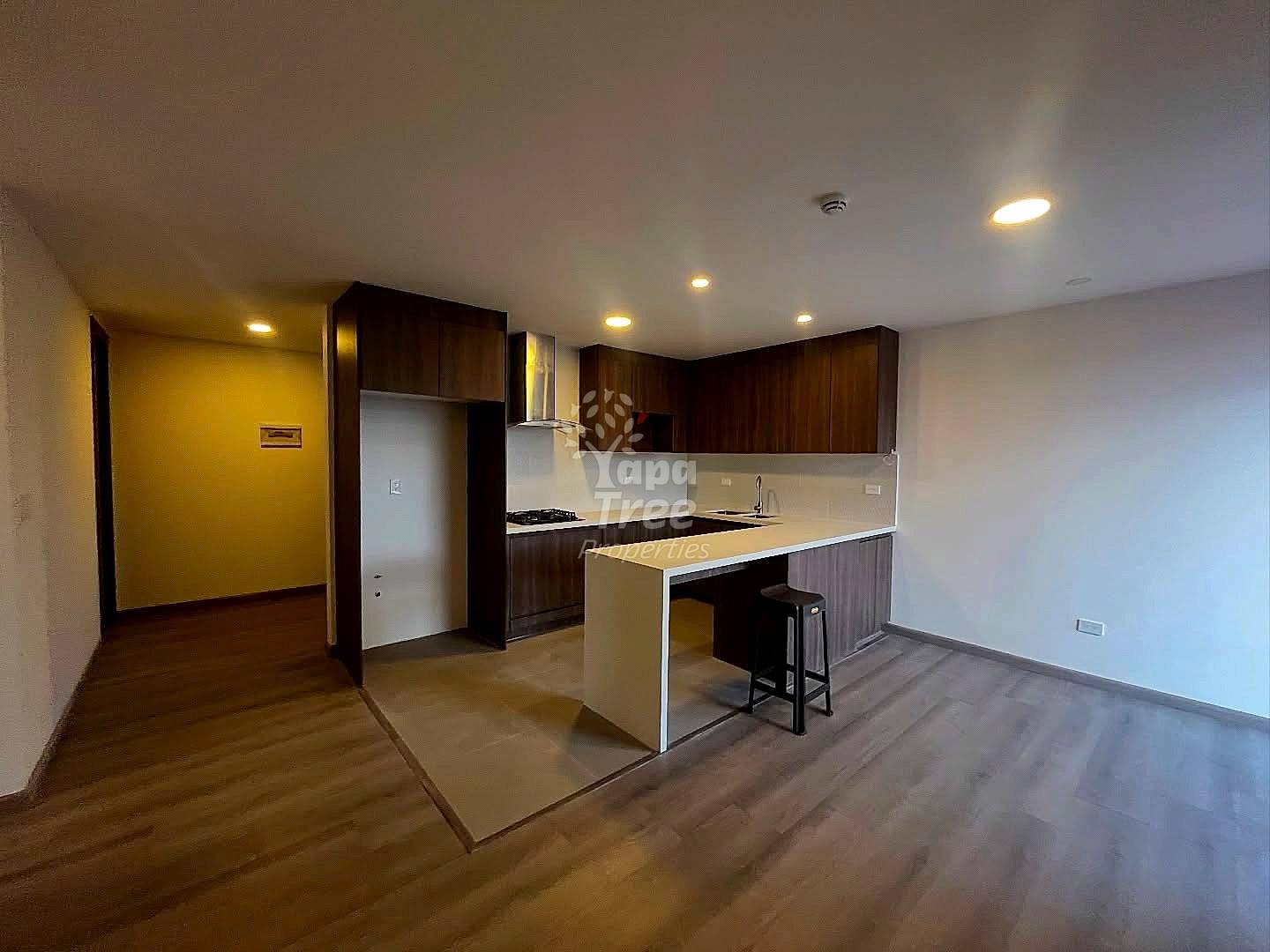 Open-plan departamento with dark wood kitchen cabinetry, white countertop island with bar seating, stainless steel appliances, and wood flooring throughout.
