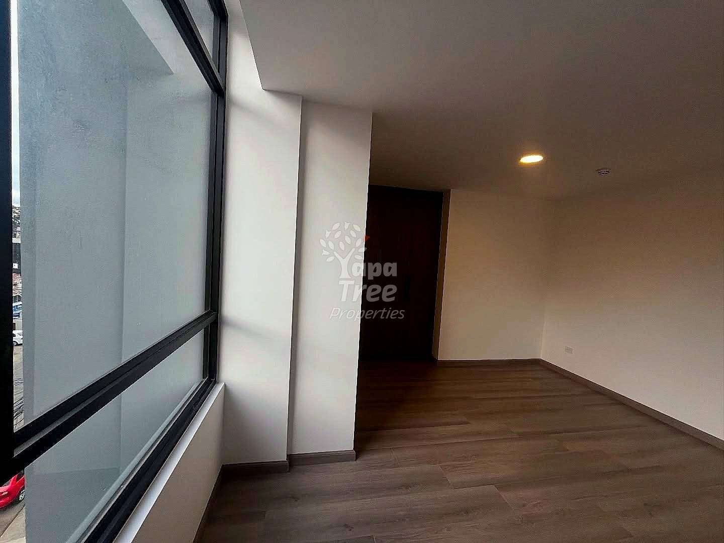 Empty room with dark wood flooring, white walls, large window with black frame, recessed ceiling light, and dark doorway entrance.